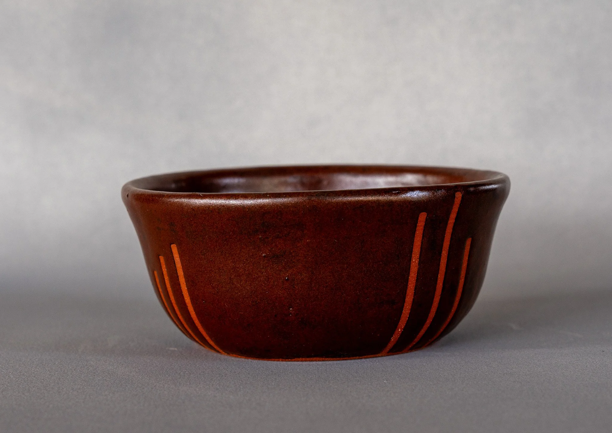 A small, round, dark brown ceramic bowl with vertical orange lines decorating the exterior, placed on a gray surface with a plain gray background.