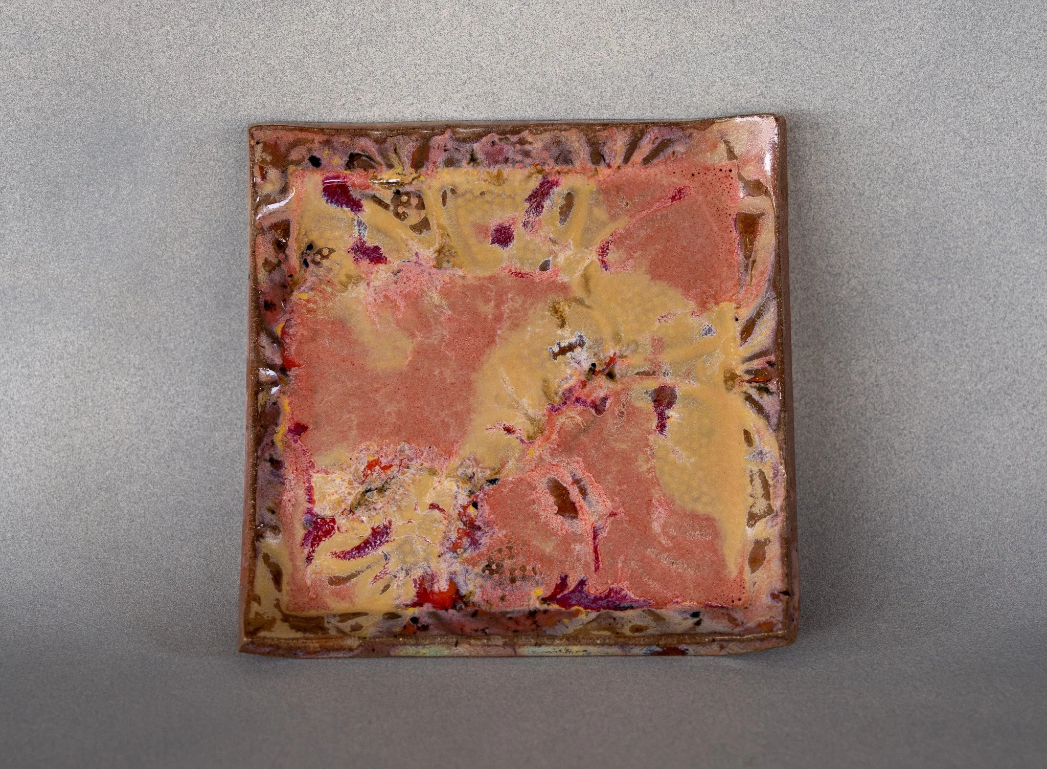 A square ceramic plate with abstract multicolored glaze pattern in shades of pink, yellow, purple, and brown