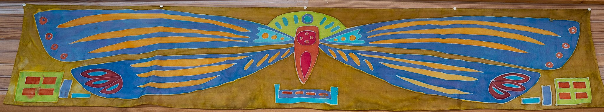 Colorful fabric banner featuring a large butterfly with blue wings decorated with orange and yellow patterns, and a central red body with green, blue, and orange accents. The background of the banner is brown, and it has geometric shapes on both ends