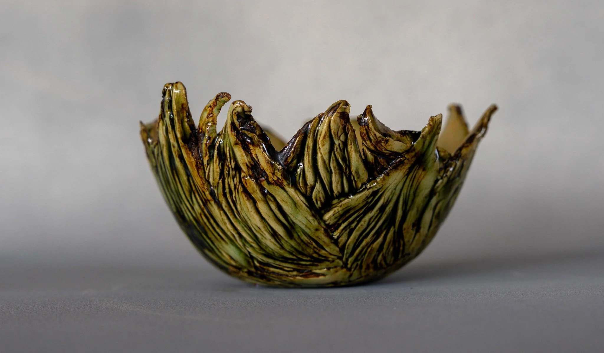 A ceramic bowl with a textured, ridged surface, glazed with green, brown, and black streaks, sitting on a gray surface against a plain gray background.