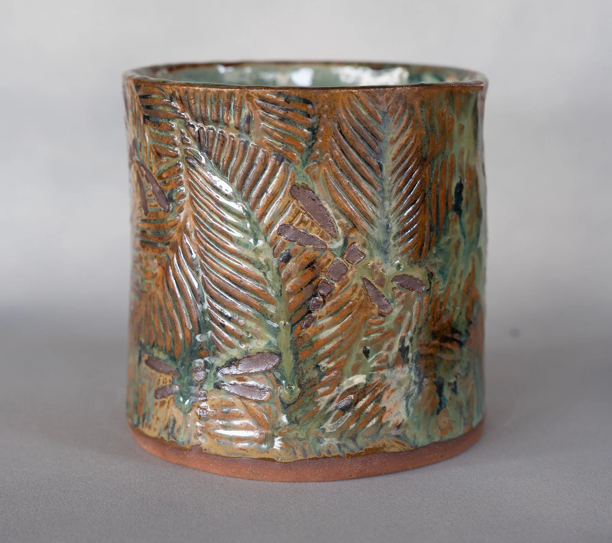 A ceramic cup with a textured surface of embedded fern leaves and plant patterns, glazed with earth-tone colors.