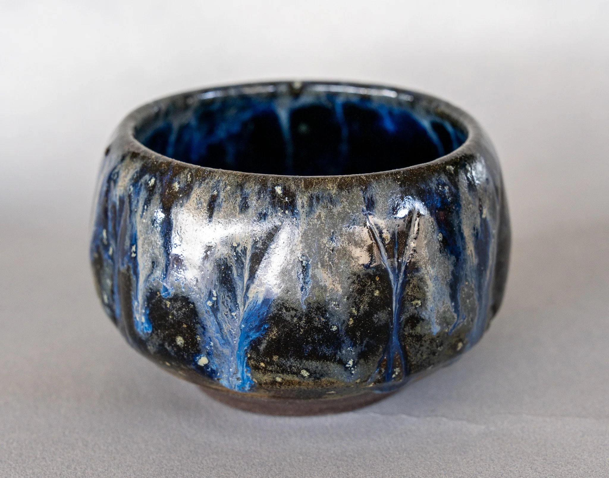 A handcrafted ceramic bowl with a textured, multicolored glaze featuring blue, black, and white accents, sitting on a gray surface.