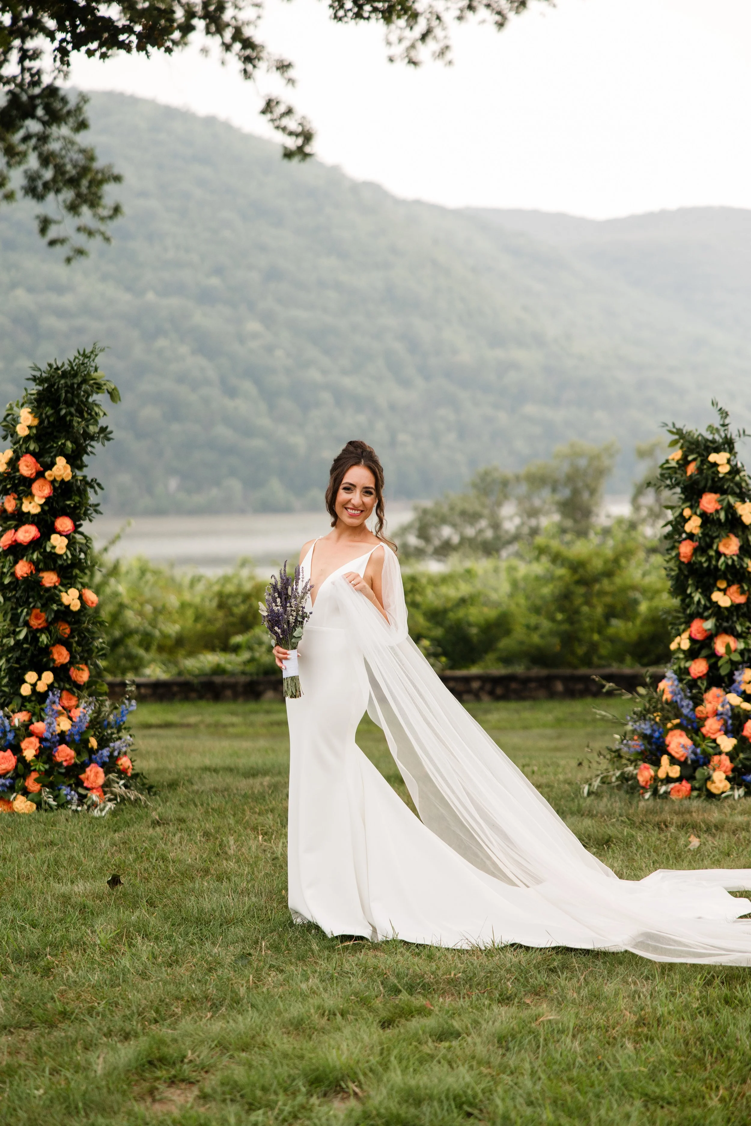 Bride in a white wedding dress holding a lavender bouquet standing outside on a grassy area with mountain scenery and floral arrangements in the background.