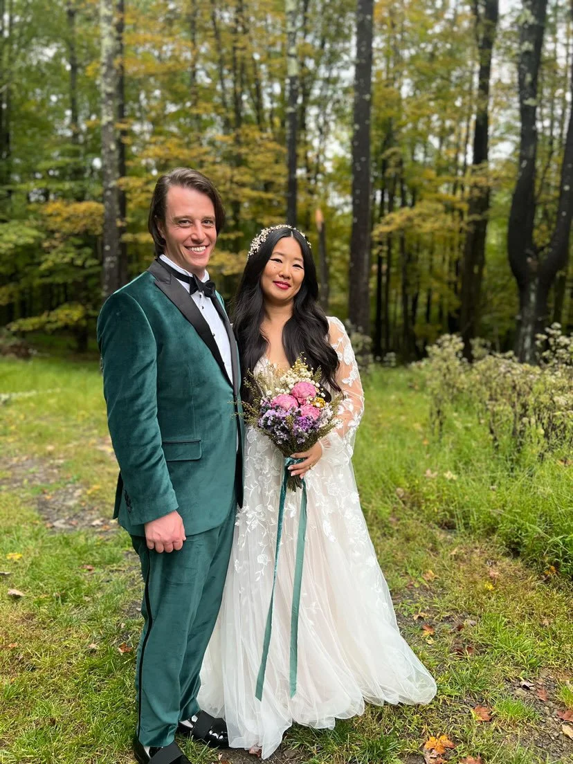A couple dressed in wedding attire standing outdoors in a forest with trees and greenery.