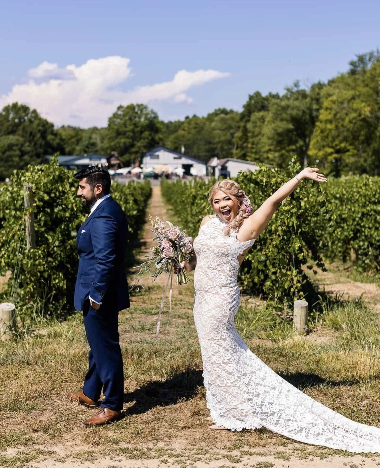 A bride in a white lace wedding dress joyfully waving, holding a bouquet, standing back to a groom in a navy blue suit with his head turned, in a vineyard on a sunny day.