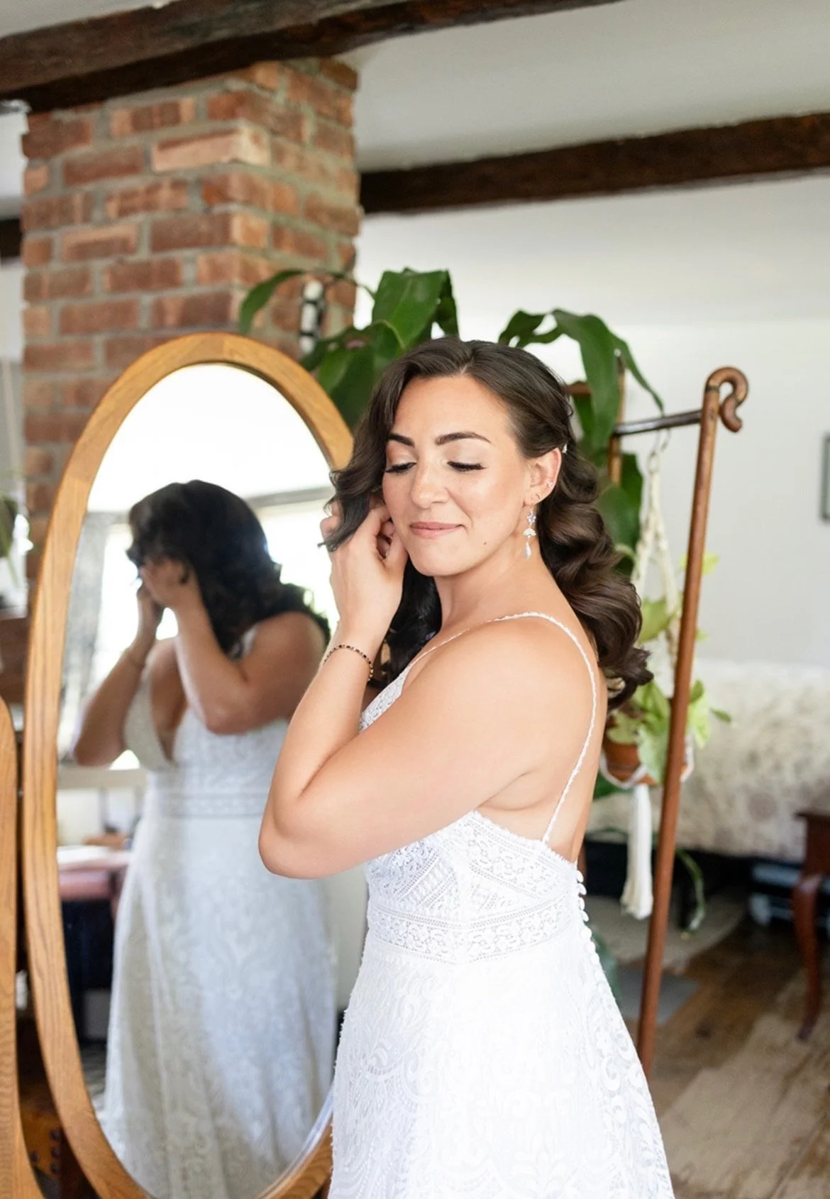 A bride in a white lace wedding dress touches her face while standing in front of a mirror, with her reflection visible. She has dark, styled hair and is wearing earrings. The background includes a brick pillar and green plants in a cozy interior setting.