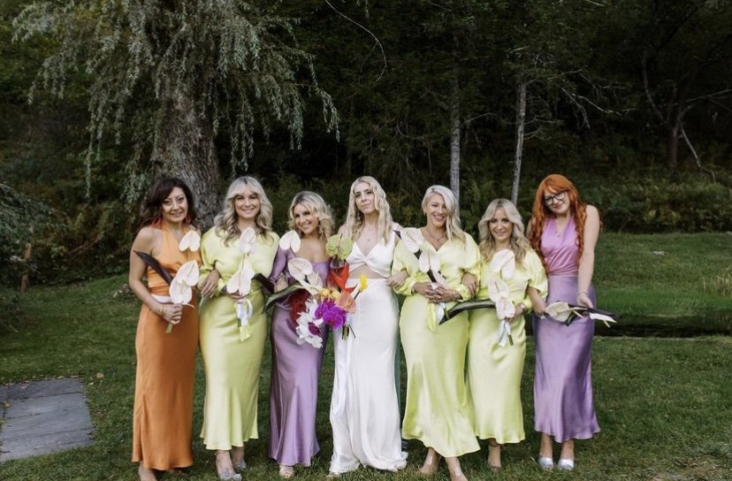 Seven women standing outdoors in a garden, dressed in colorful dresses and holding white calla lilies.