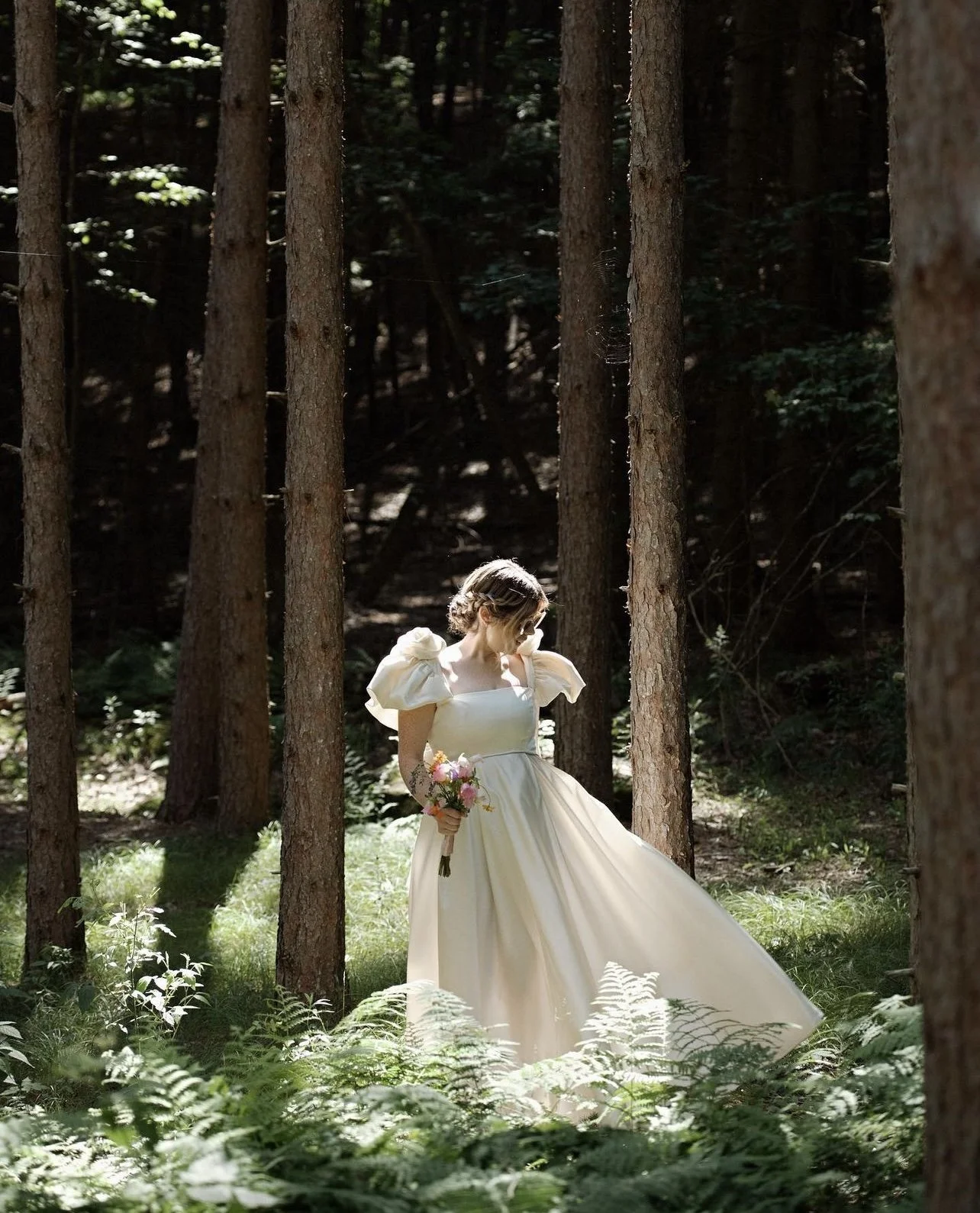 A woman in a white dress holding a bouquet of flowers standing in a forest with tall trees and green ferns.