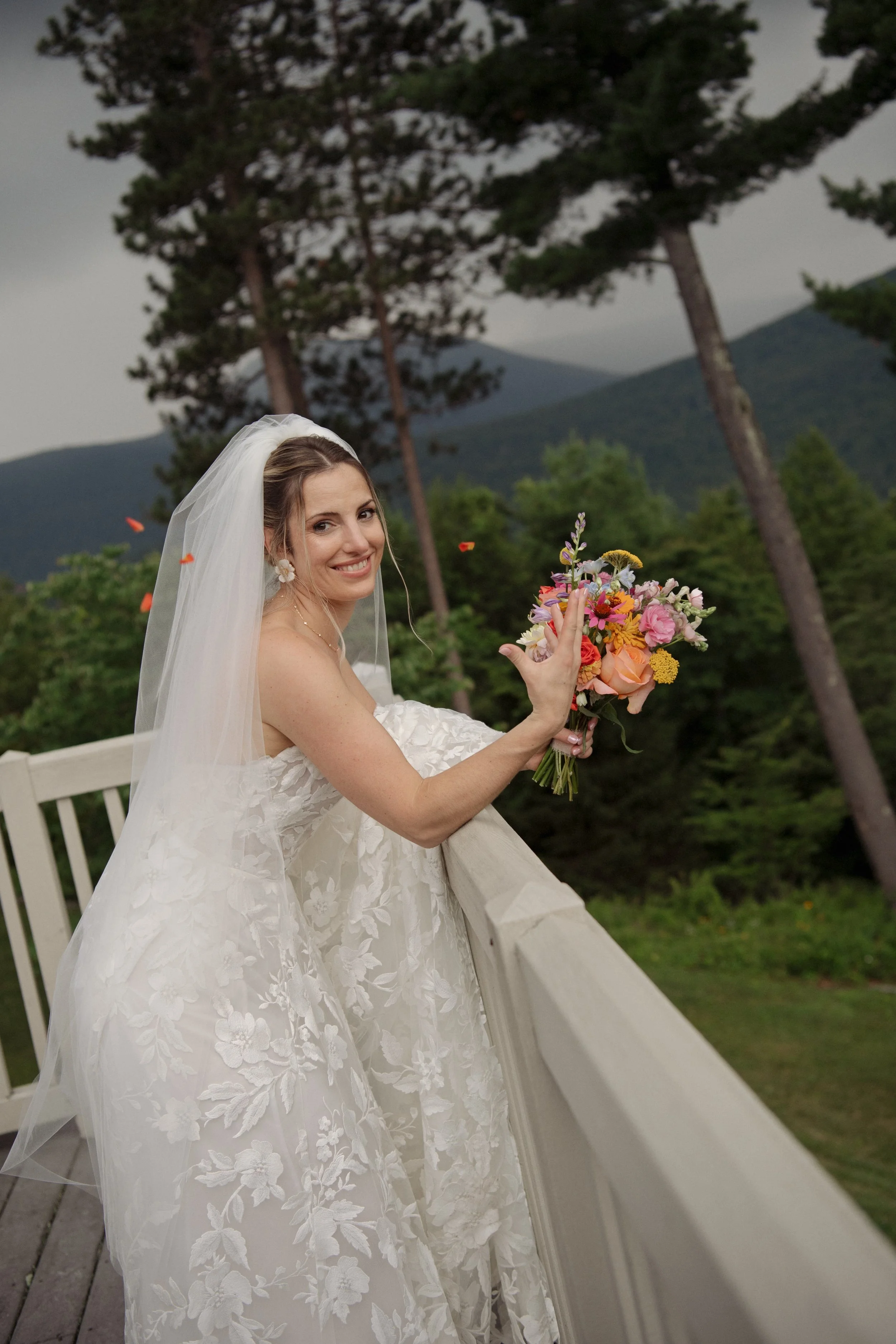 A smiling bride in a white lace wedding dress and veil holding a colorful bouquet of flowers on a balcony with trees and mountains in the background.