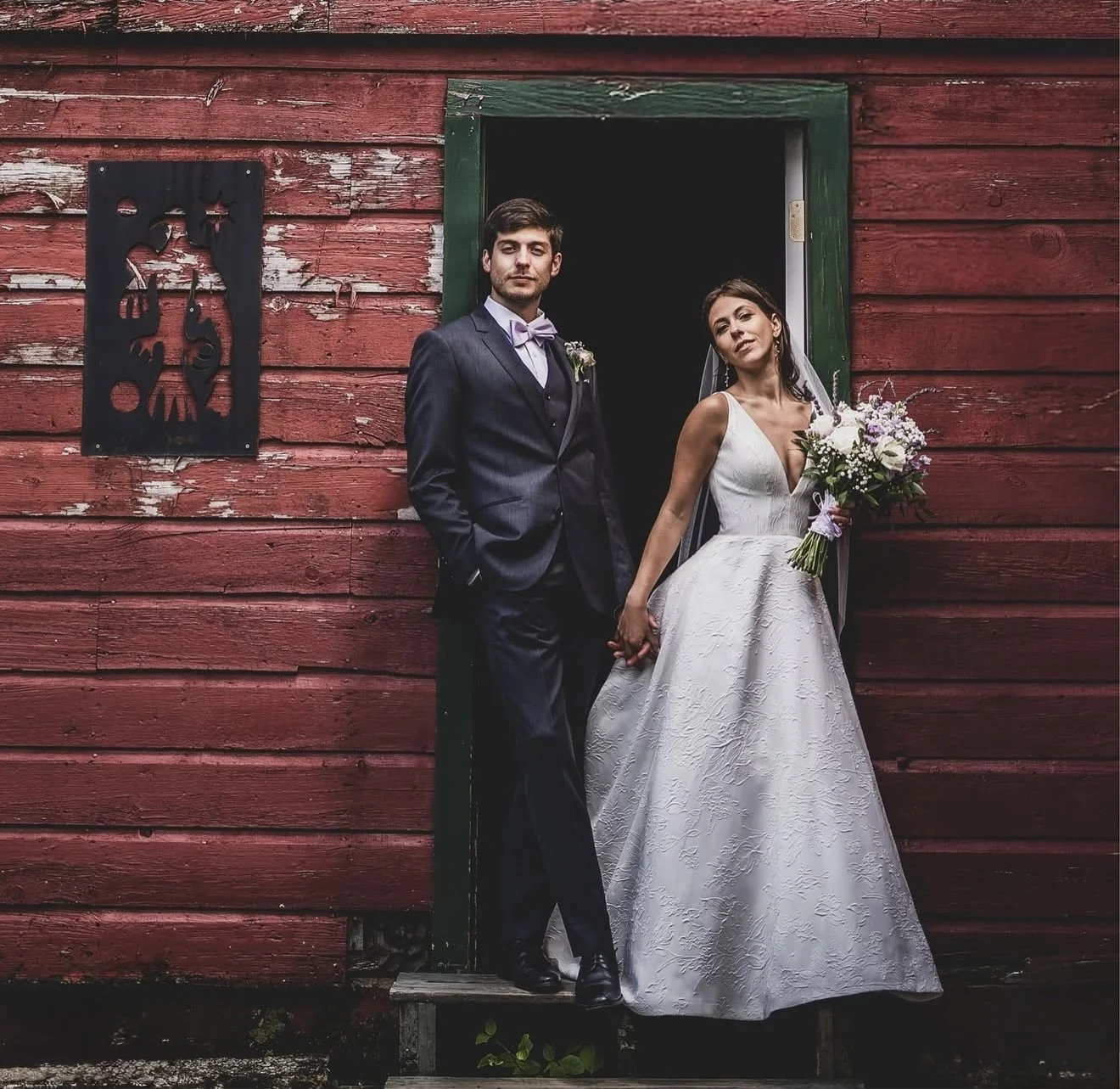 A bride and groom standing hand in hand outside a rustic red barn, dressed in wedding attire. The groom is in a dark suit with a bow tie, and the bride in a white gown holding a bouquet of white and purple flowers.