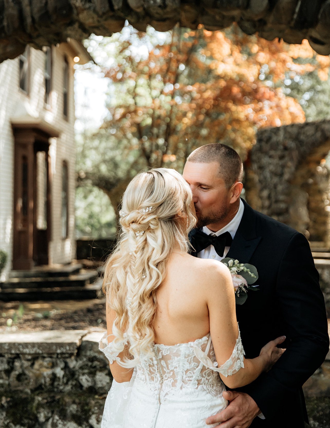 A bride and groom sharing a kiss outdoors, with autumn trees and a rustic building in the background.