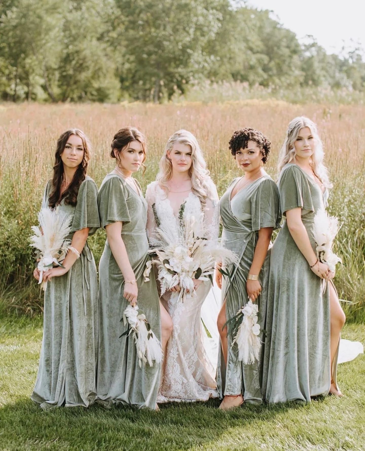 A group of five women standing outdoors in a grassy field with trees in the background, dressed in matching green velvet dresses, holding bouquets, with the woman in the middle wearing a white lace dress.