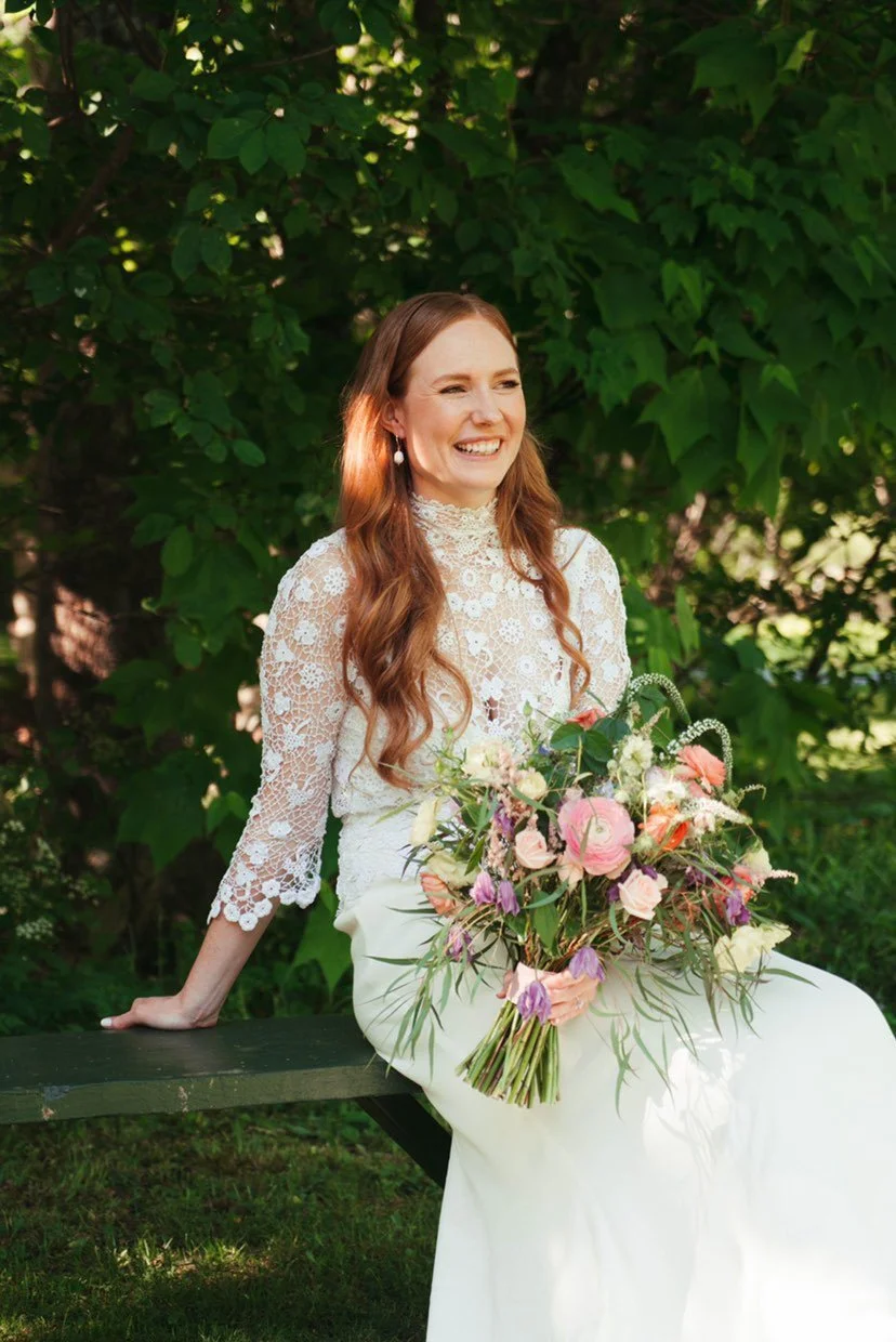 A woman wearing a lace wedding dress with long, wavy reddish-brown hair, sitting on a green bench outdoors, holding a bouquet of pink, purple, and white flowers, and smiling.