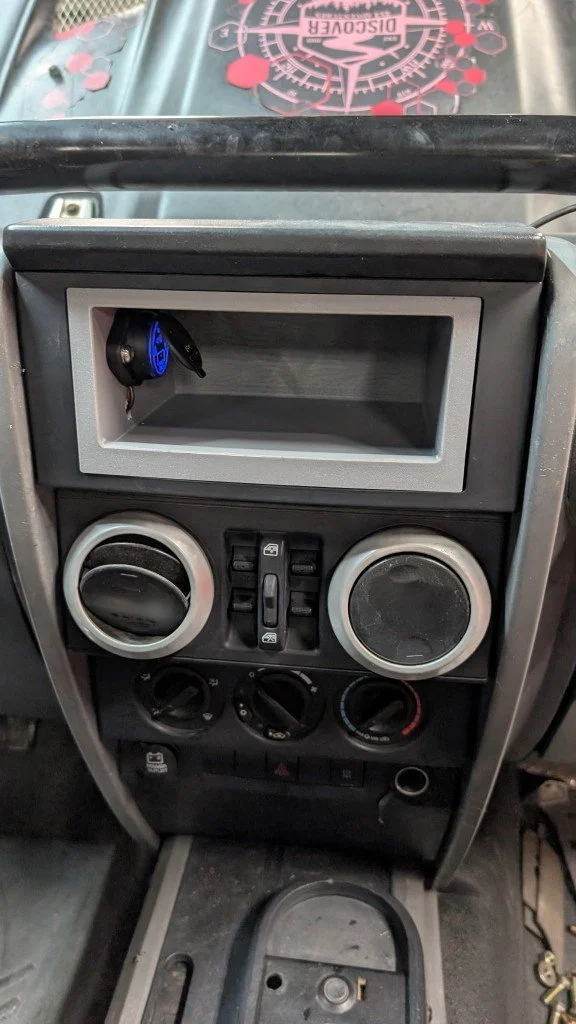 The image shows a vehicle's dashboard with a section for a stereo or GPS unit, which is currently empty. Below are two round air conditioning vents, a set of control switches, and climate control dials. There is a small storage compartment with a power outlet or charging port at the bottom.
