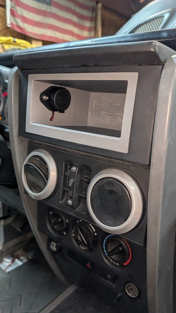 The image shows the dashboard of a vehicle, featuring air conditioning controls, a small open storage compartment with a black electrical device inside, and various switches and knobs.