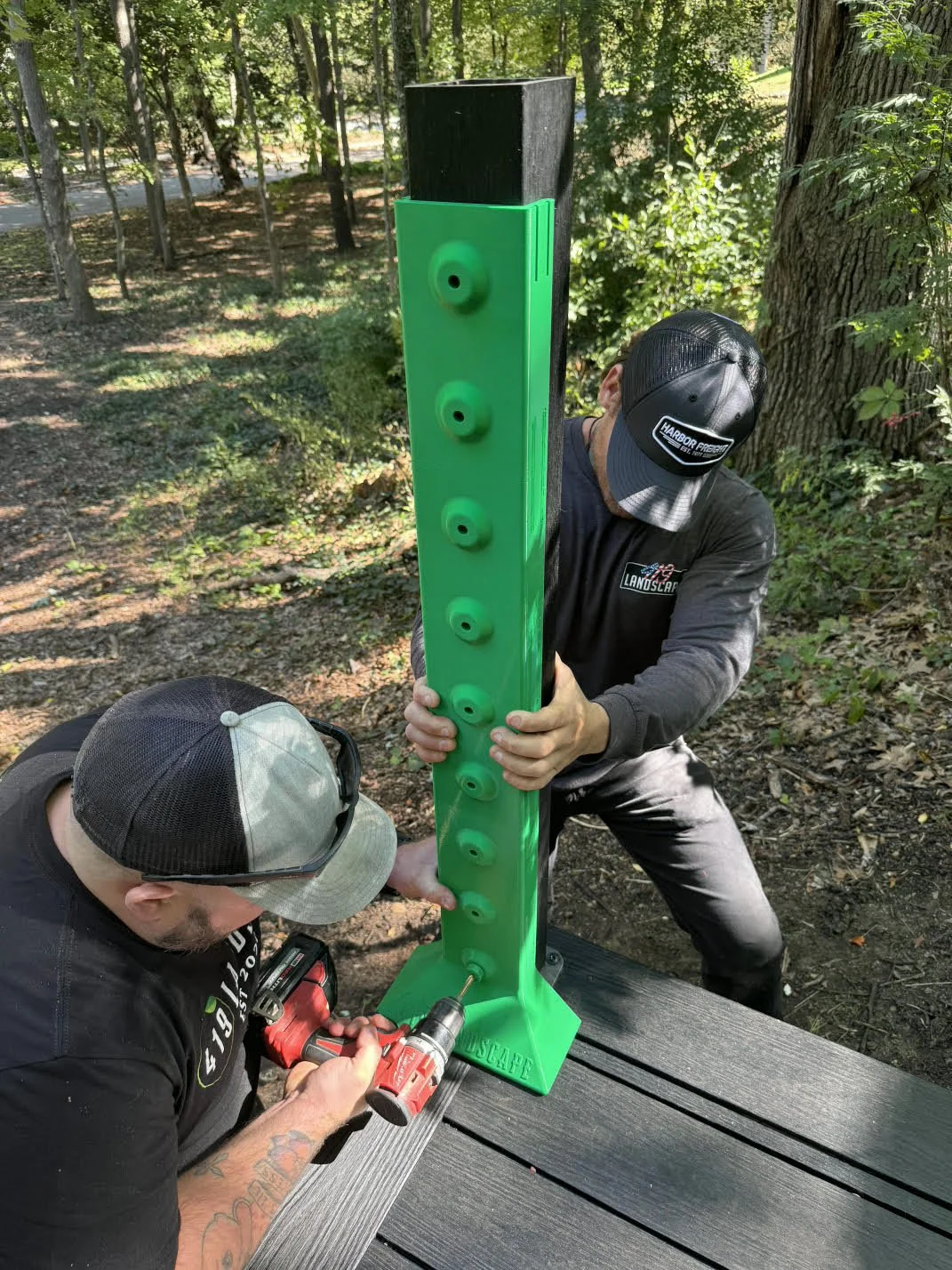 Two men assembling a large green plastic obstacle with footholds outdoors in a wooded area.