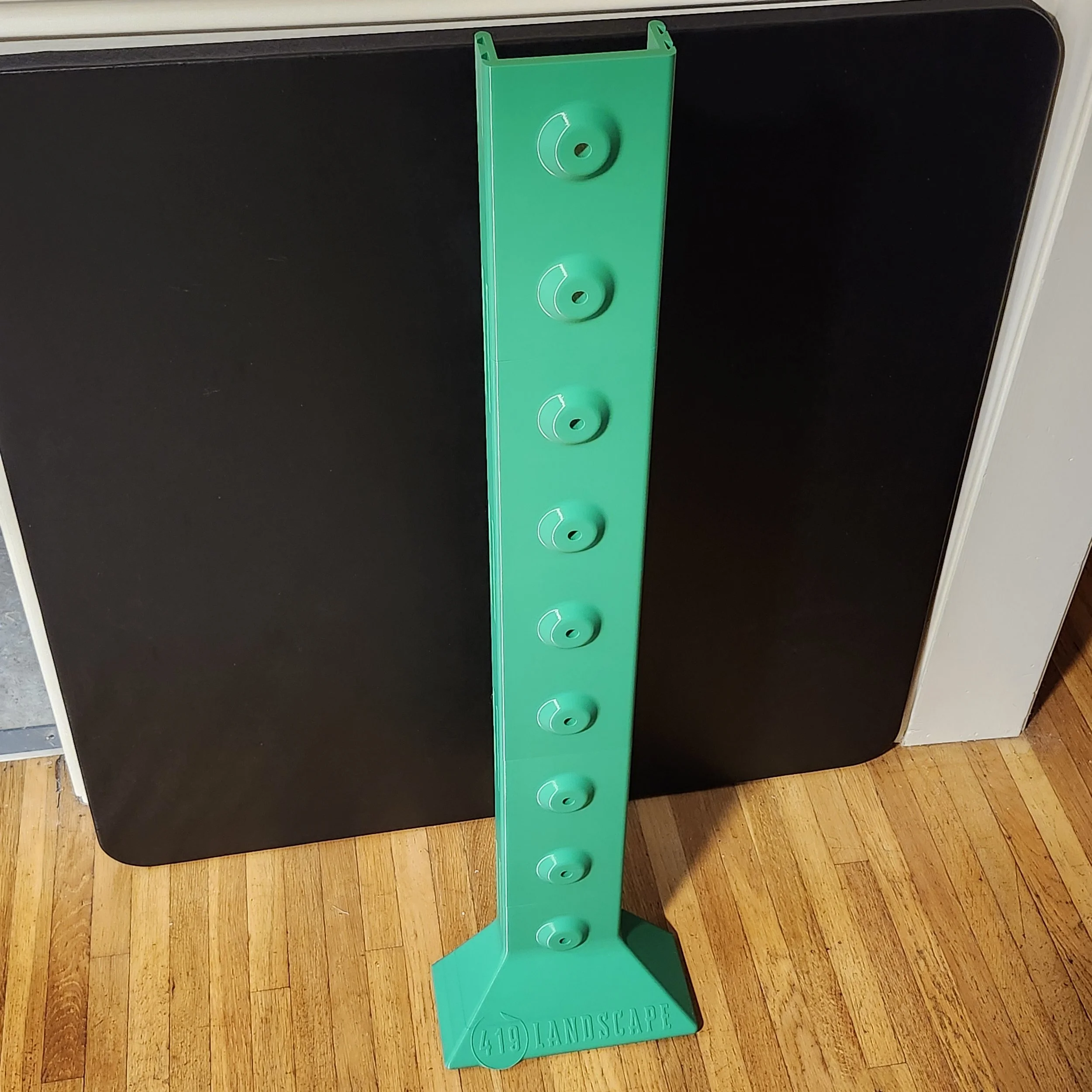 A green landscape utility rack with four circular holes, standing on a wooden floor in front of a black appliance.