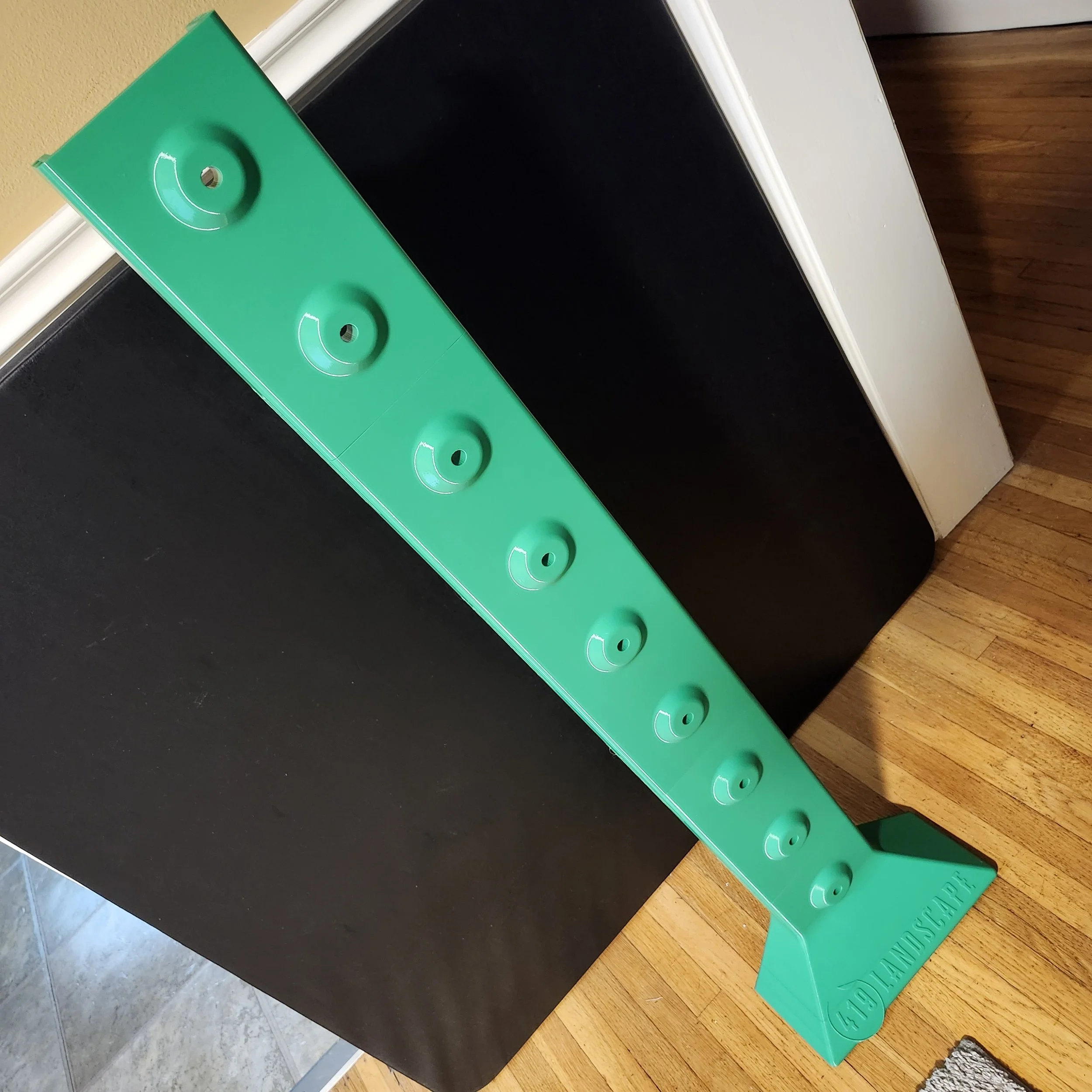 A green plastic tool has a curved design with circular cutouts and is placed on top of a black object, on a wooden surface. The background includes a beige wall and a section of tiled floor.