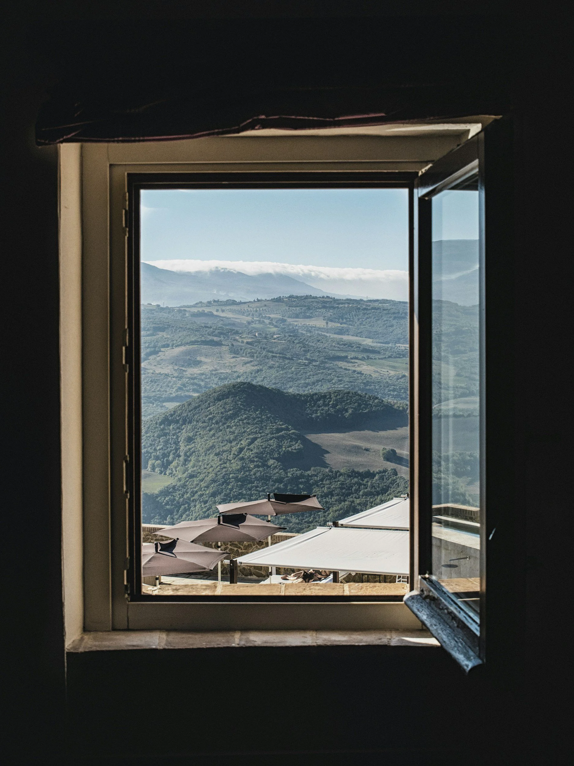 View of a landscape with rolling hills, mountains in the distance, and a cloudy sky, seen through a window with open shutters.