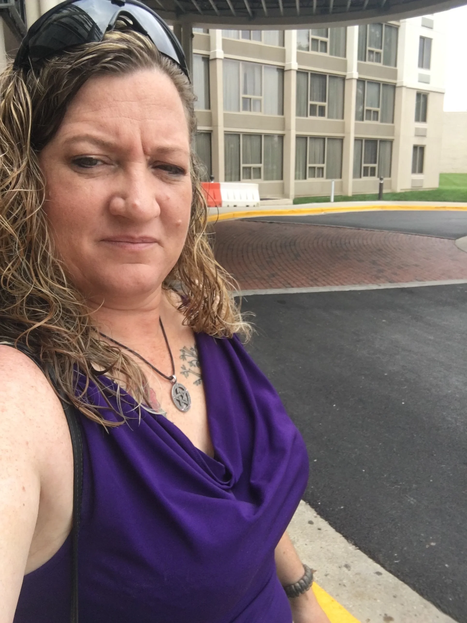 A woman with curly hair and wearing a purple top with a necklace, taking a selfie outdoors near a building with multiple windows, on a sidewalk with a brick-paved street and a yellow curb.