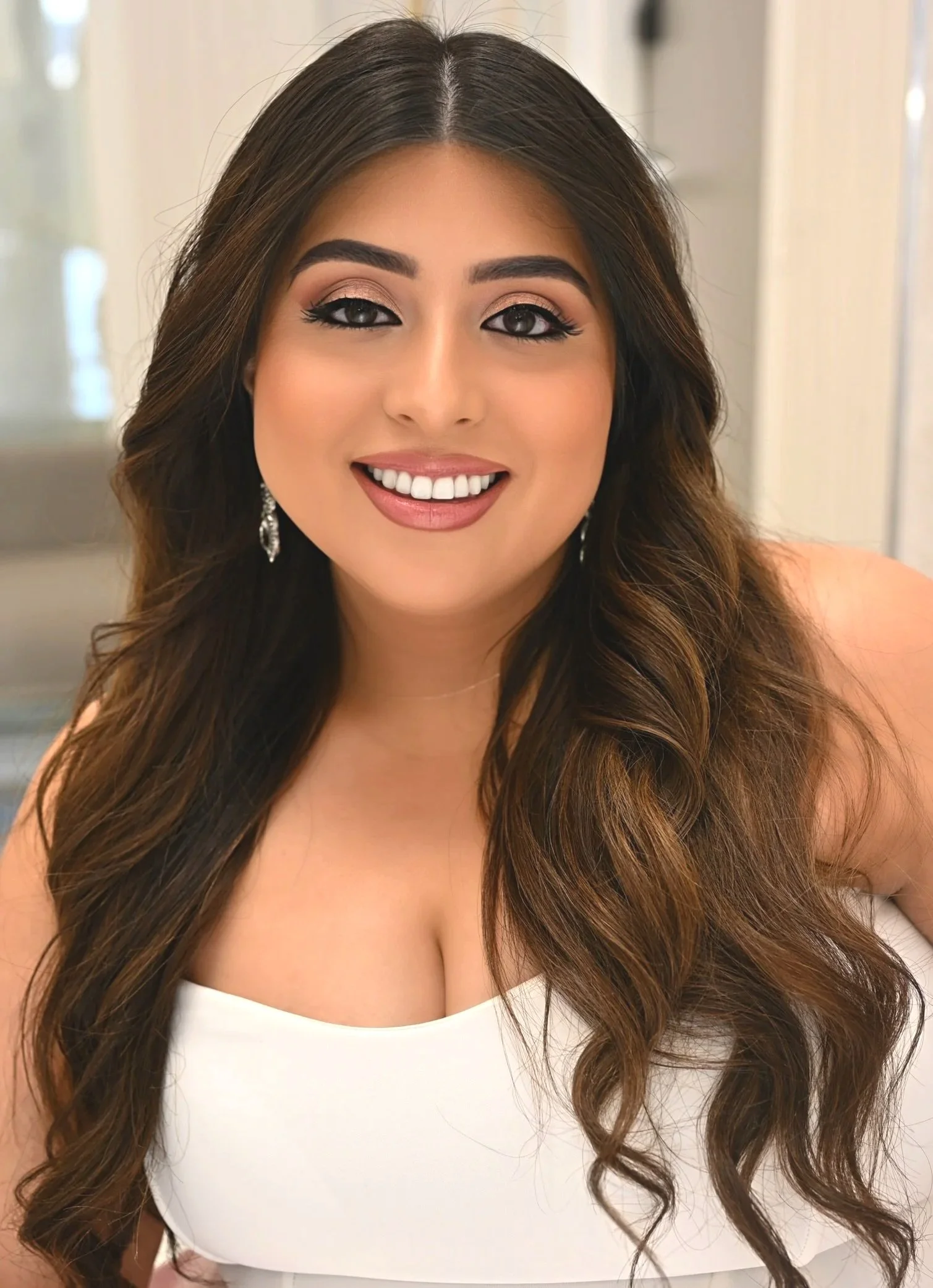 Close-up of a smiling bride with long, wavy brown hair, wearing makeup, silver earrings, and a white top.