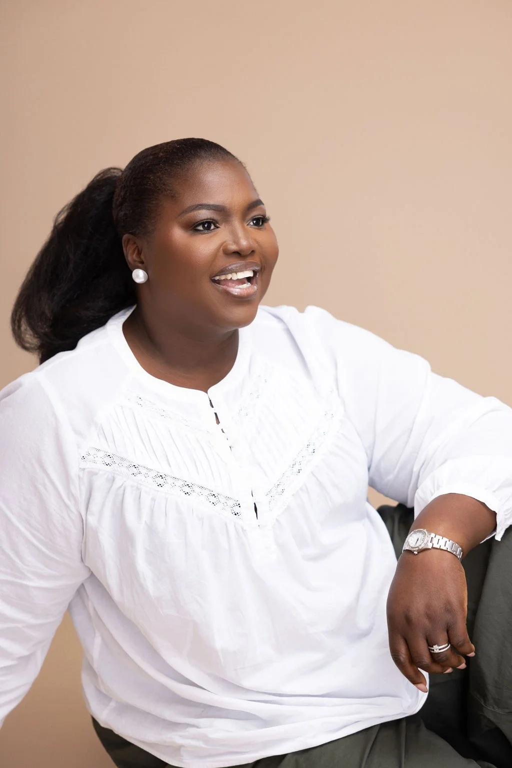 A woman with dark hair tied back, wearing a white blouse, pearl earrings, a watch, and a ring, smiling and sitting with her arm resting on her knee against a beige background.