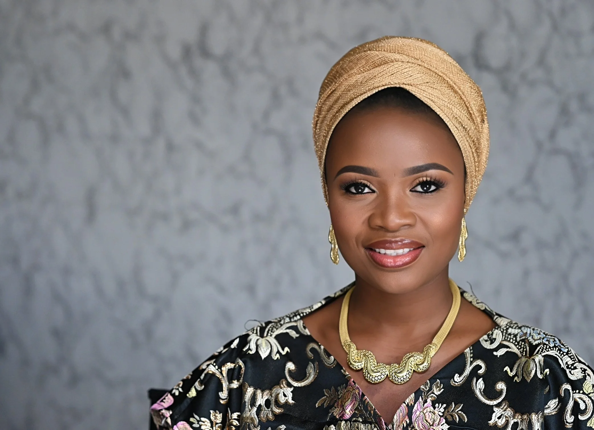 A woman of African descent with a warm smile wearing a gold headwrap, gold jewelry, and a floral-patterned dress, standing against a neutral gray textured background.