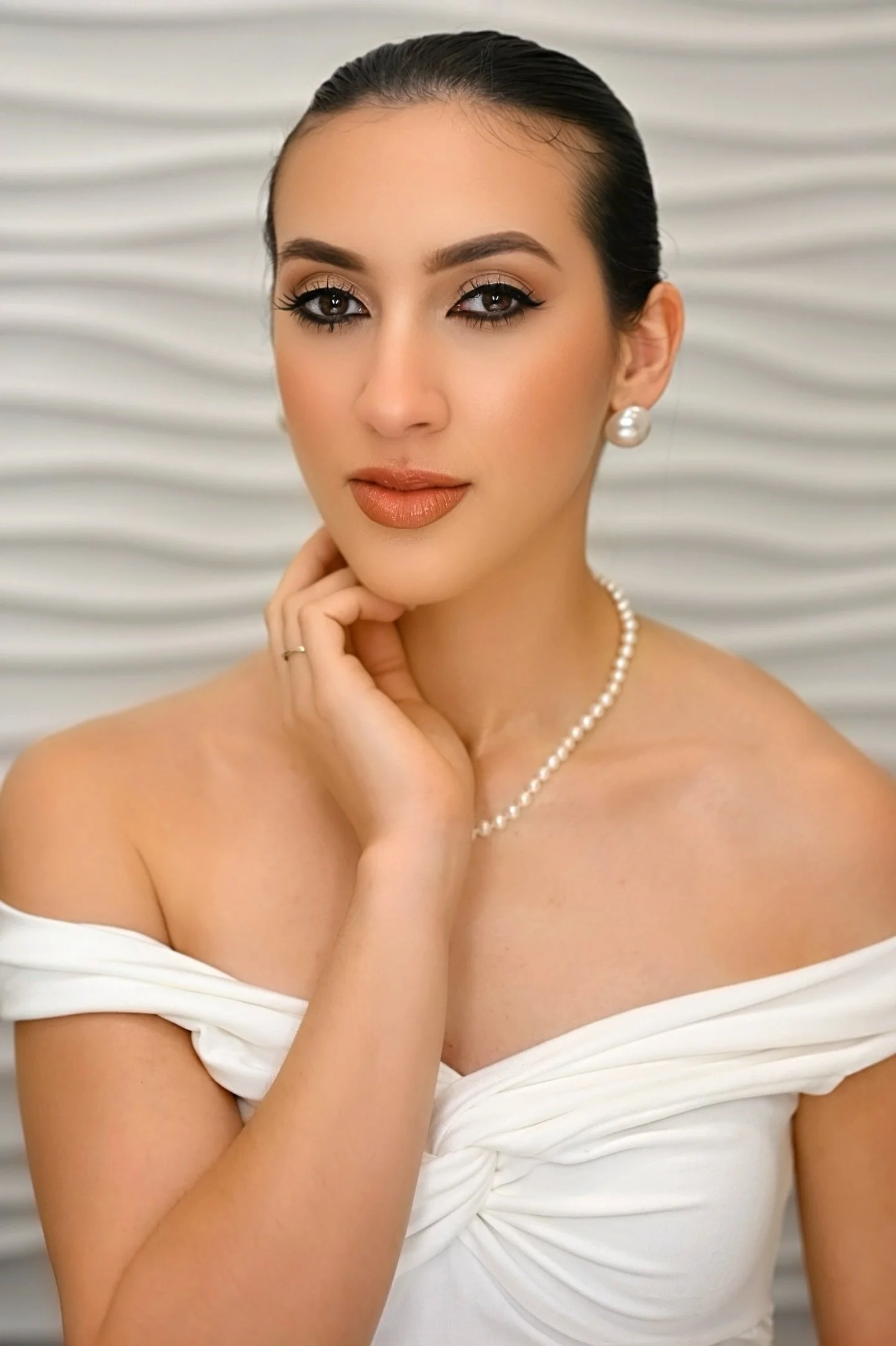 Close-up of a woman with dark hair in a bun wearing pearl earrings and necklace, and an off-shoulder white dress, with a textured gray background.