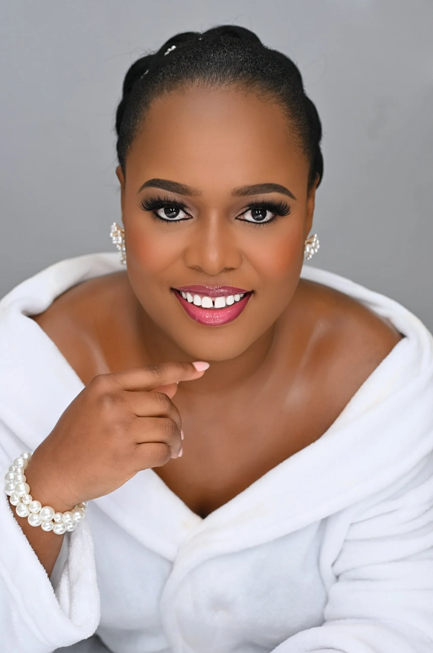 A bride with dark hair styled in an elegant updo, wearing pearl earrings and bracelet, smiling with white teeth, makeup, and a white towel draped around her shoulders against a plain gray background.