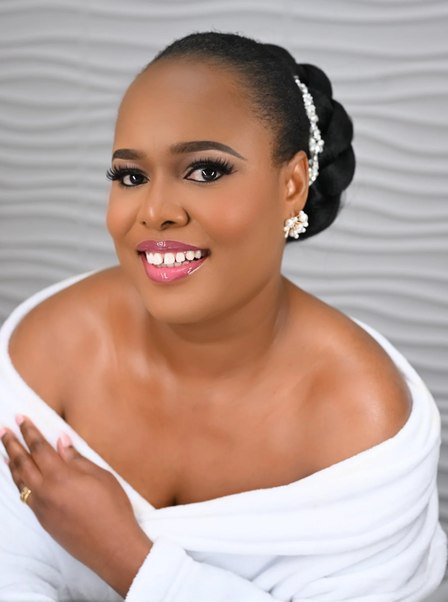 Bride with dark hair styled in an elegant updo, wearing pearl earrings, and a white towel wrapped around her shoulders, smiling warmly against a white textured background.