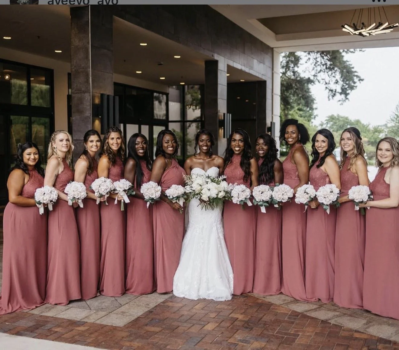 Bride in a strapless white wedding gown surrounded by bridesmaids in mauve dresses, holding white floral bouquets outside a modern venue.