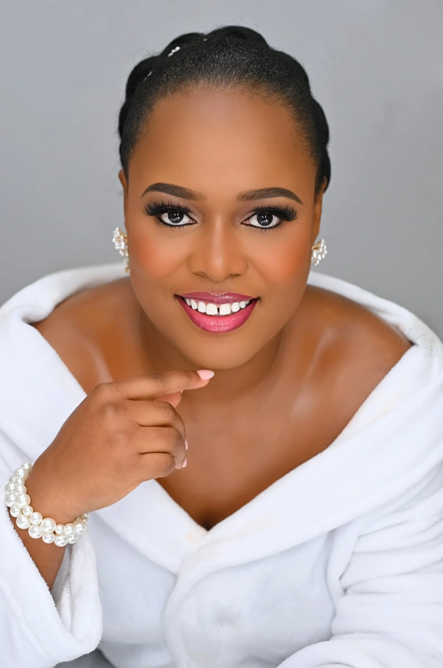 A woman with a warm smile, wearing big earrings and a pearl bracelet, wrapped in a white towel, with her hand near her chin, against a neutral gray background.