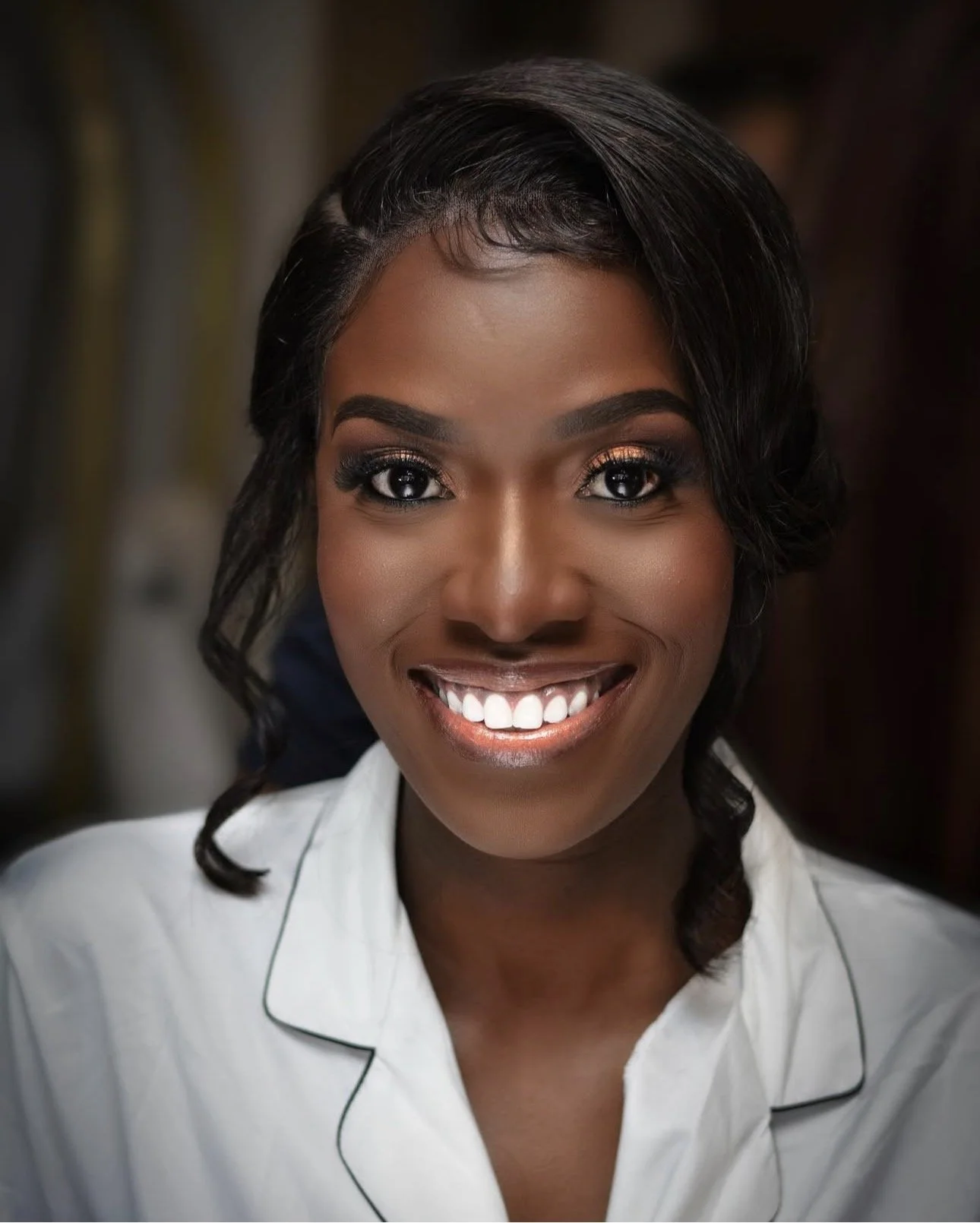 Close-up portrait of a smiling African American woman with dark wavy hair, wearing a white collared shirt with black piping, and makeup including bold eye makeup and lipstick.