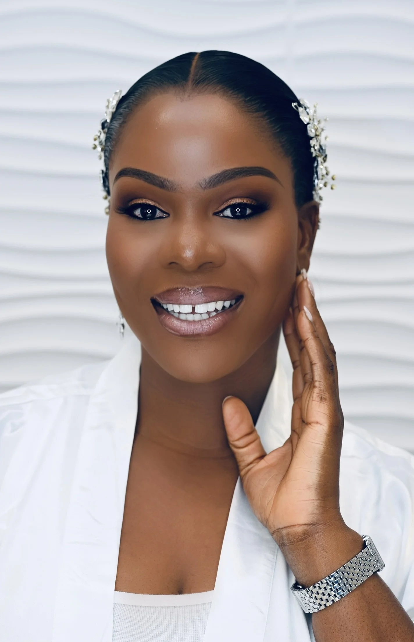 Close-up portrait of a smiling bride with dark hair styled back, wearing pearl hair accessories and a white outfit, against a textured white background.