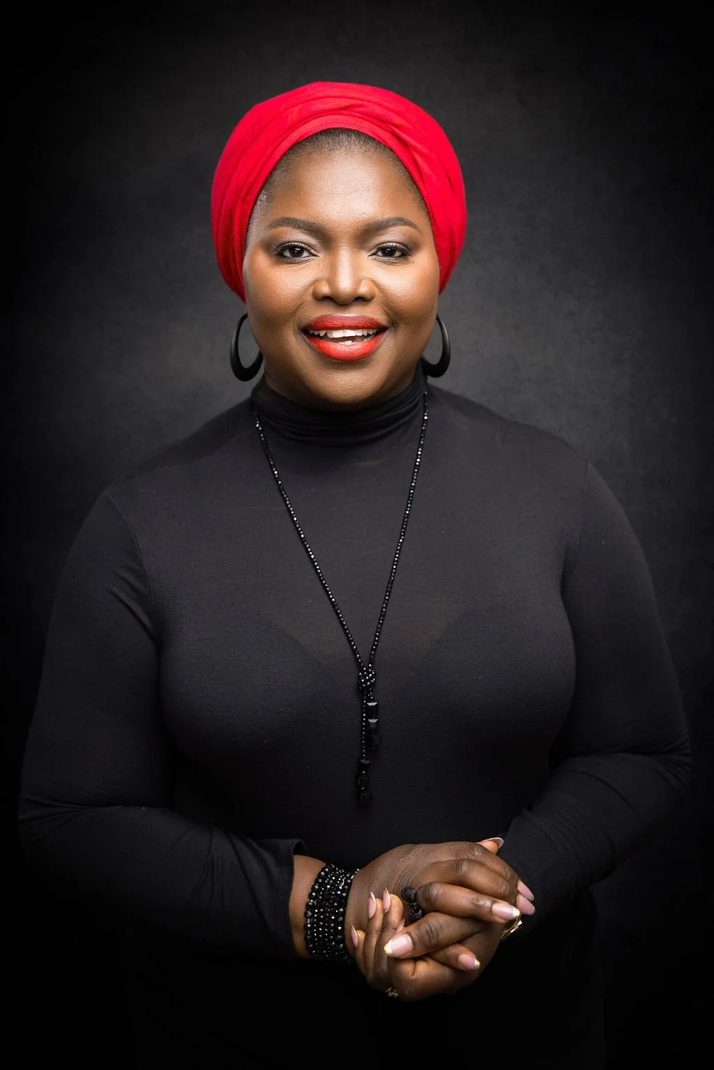 Portrait of a woman wearing a red headscarf, black turtleneck, and jewelry, smiling against a dark background.