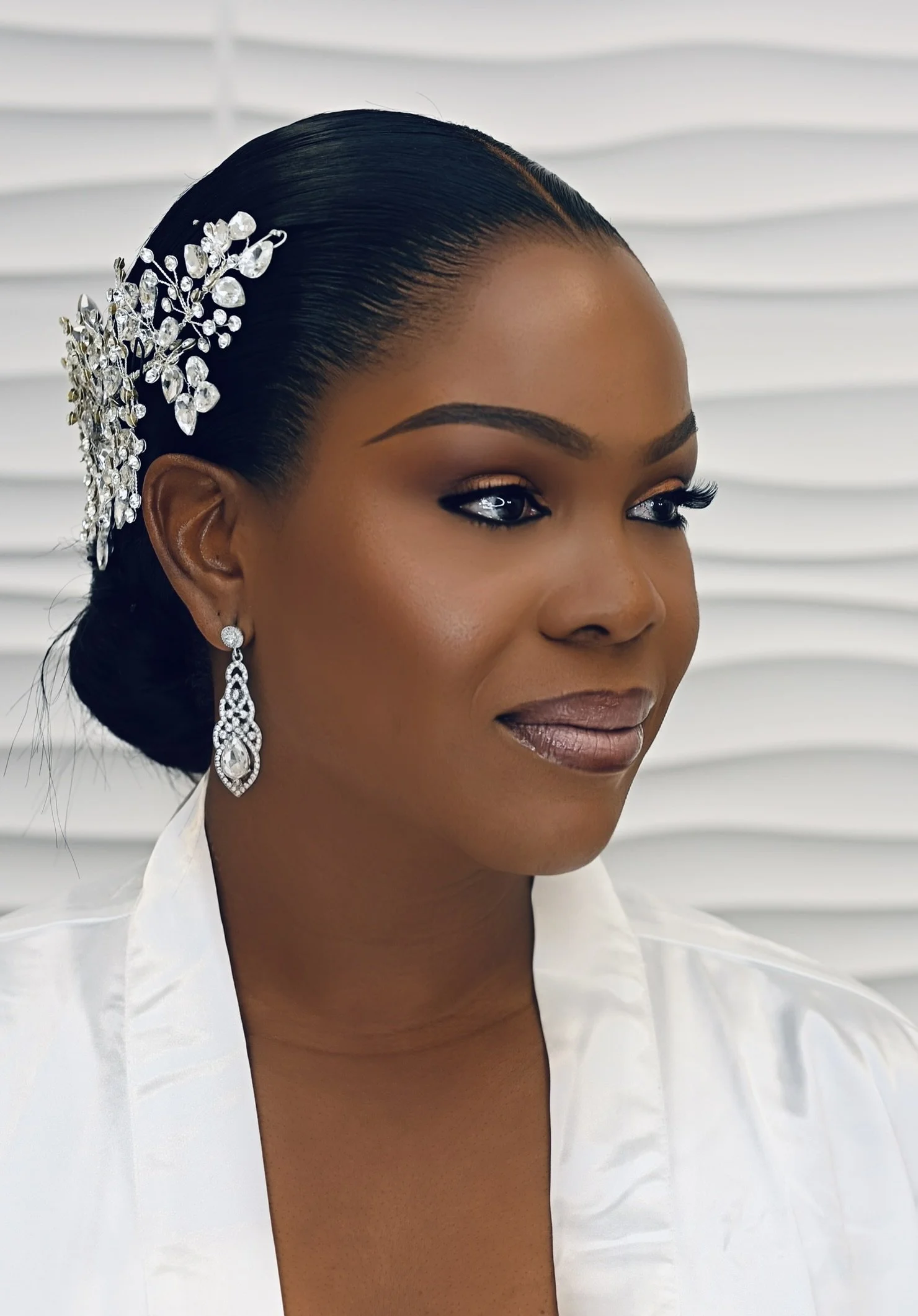 Close-up portrait of a woman with a sleek, pushed-back hairstyle adorned with a rhinestone hairpiece, wearing elegant drop earrings, and a white satin blouse, standing in front of white window blinds.