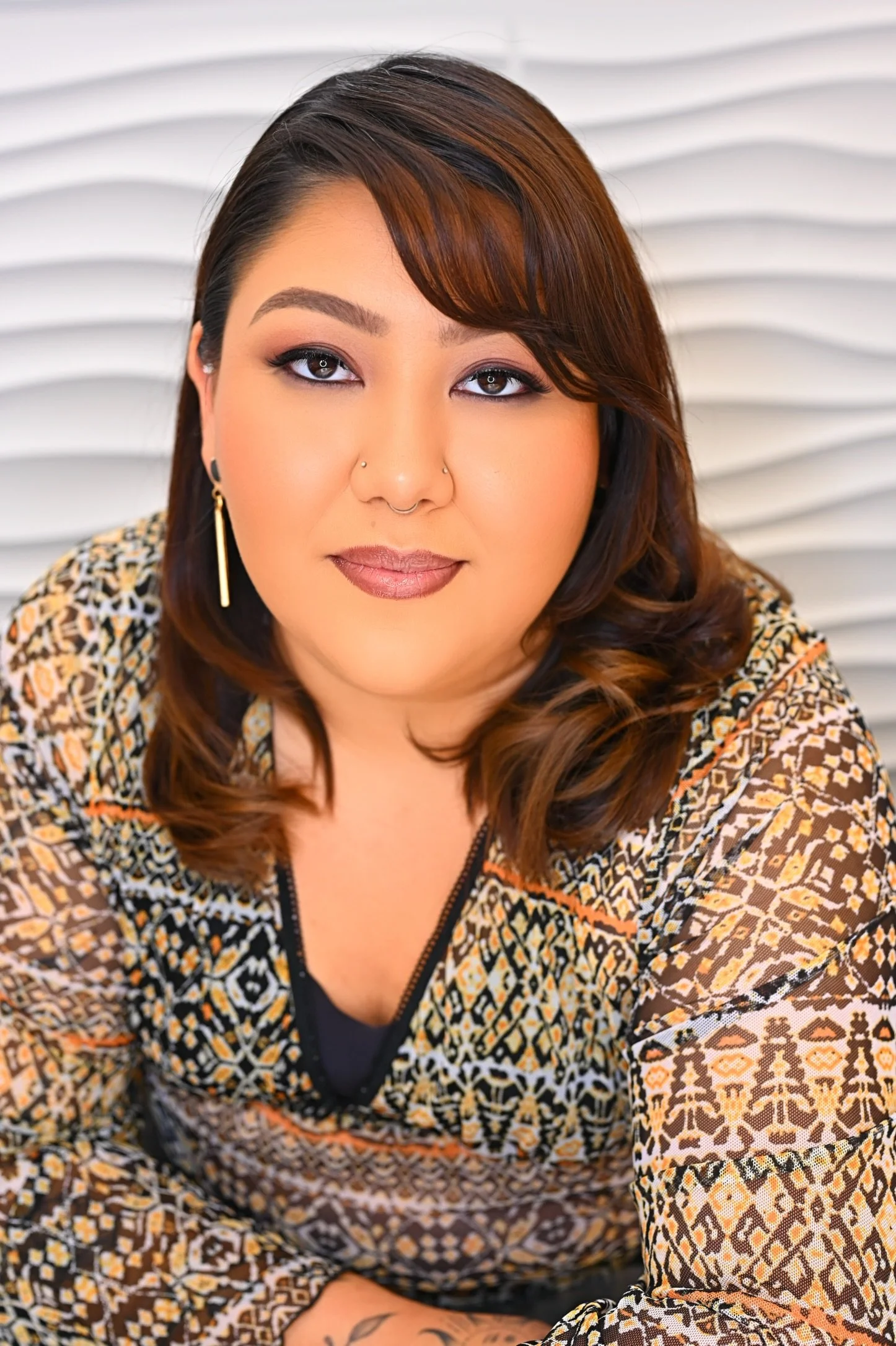 A woman with brown hair, tan skin, and makeup, wearing earrings and a patterned top, sitting in front of a white textured background.