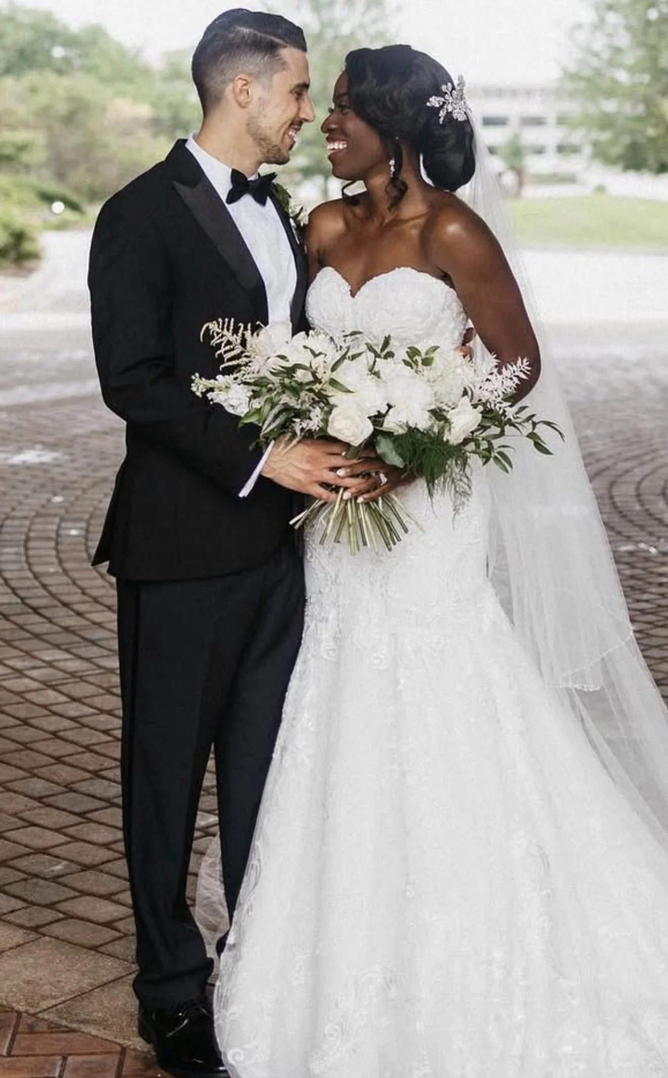 Bride in a strapless lace wedding gown holding a white floral bouquet, standing with groom in a black tuxedo during an outdoor wedding portrait.