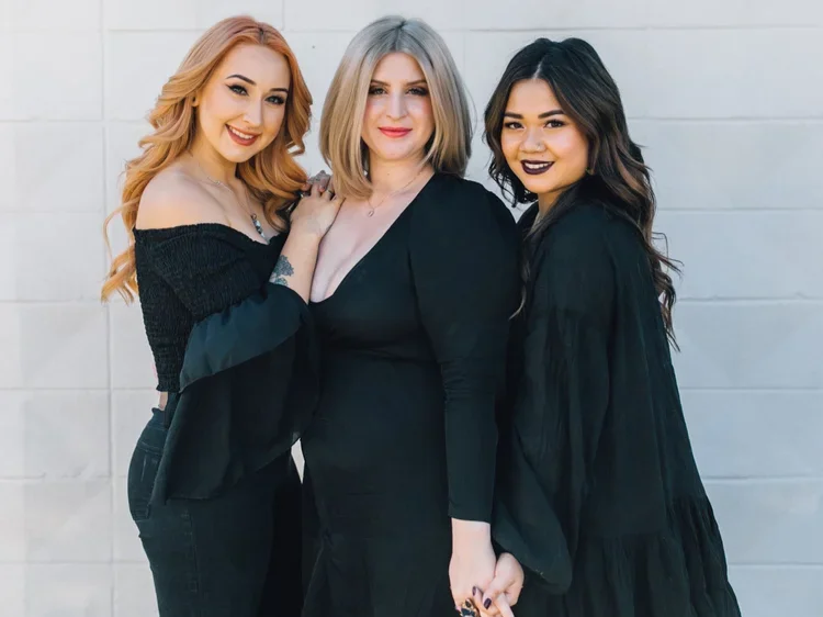 Three women standing together against a white wall, smiling, with one woman in the center holding her hand to her side, they are dressed in dark clothing.
