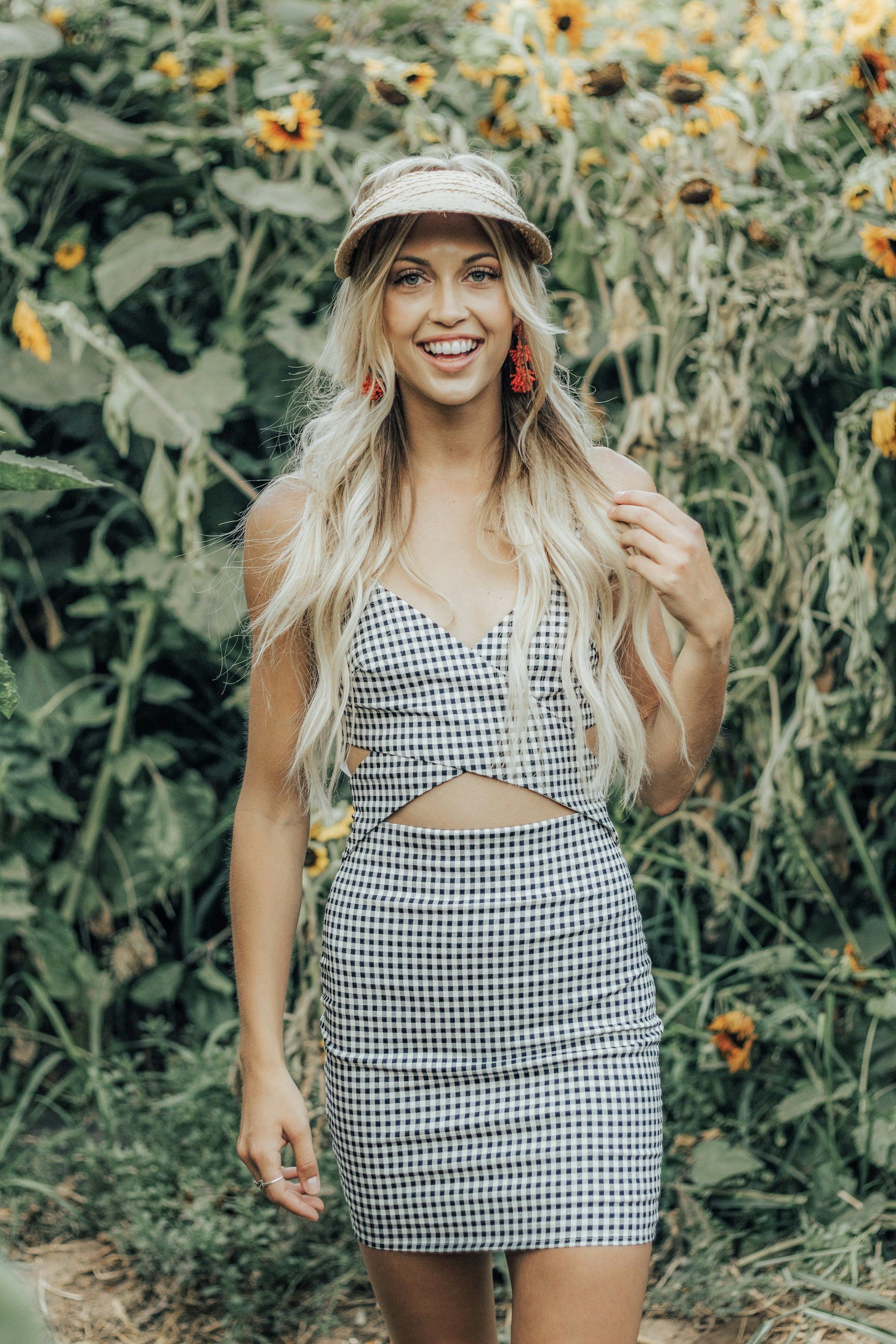 A young woman with long blonde hair, smiling and showing her tongue, wearing a checkered dress with cutouts and a visor hat, standing outdoors in a garden with sunflowers and greenery.