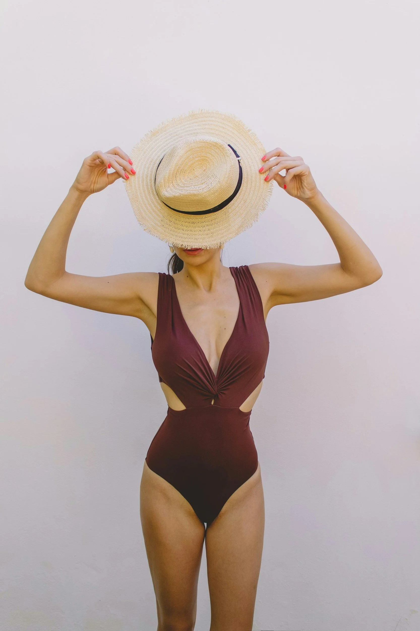A woman wearing a maroon dress with cutouts, holding a wide-brimmed straw hat in front of her face, standing against a plain white wall.