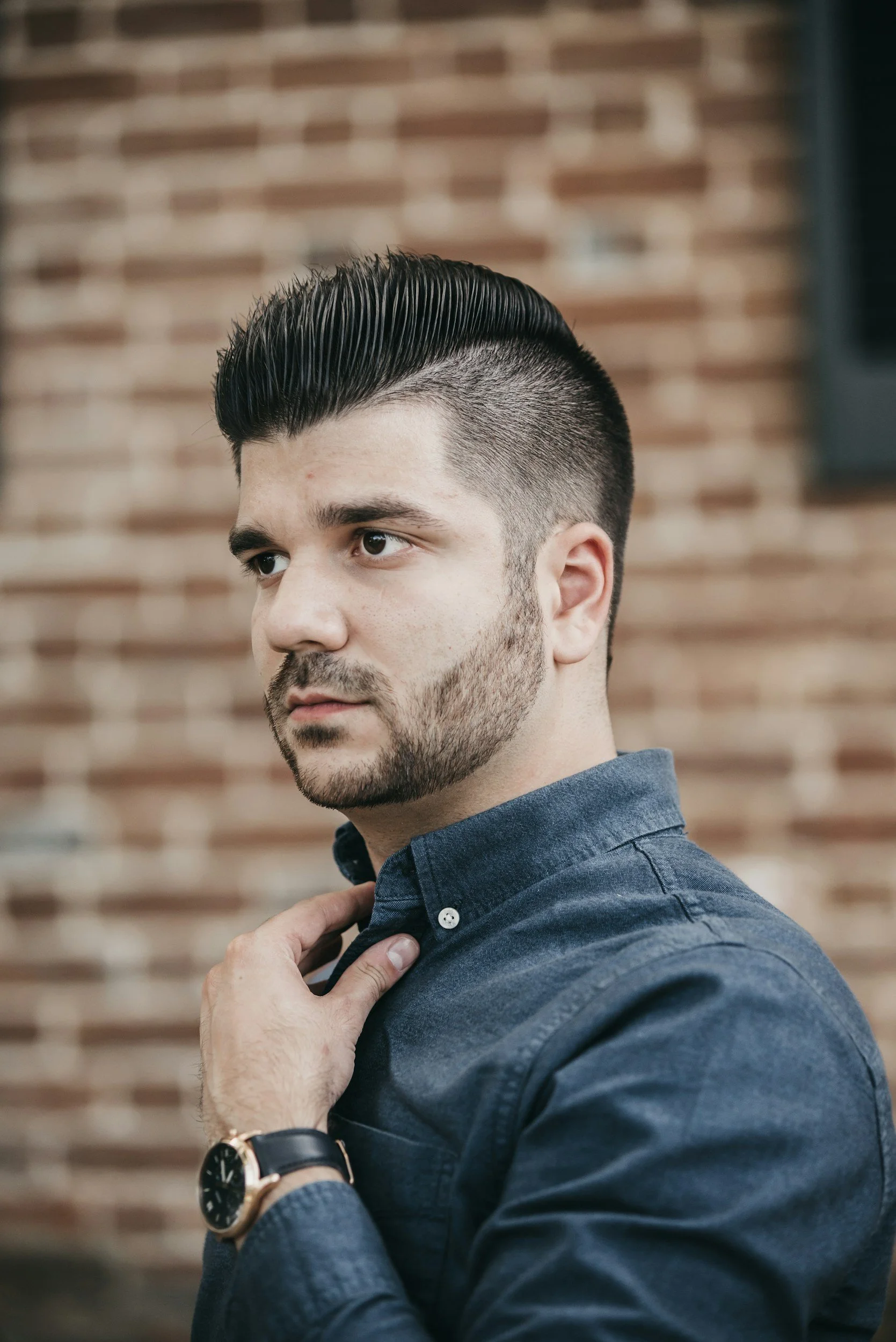 A young man with dark hair styled back, a beard, and a mustache, wearing a dark blue shirt, adjusting his collar with his left hand, and a black wristwatch with a leather strap on his left wrist. He stands in front of a brick wall background.