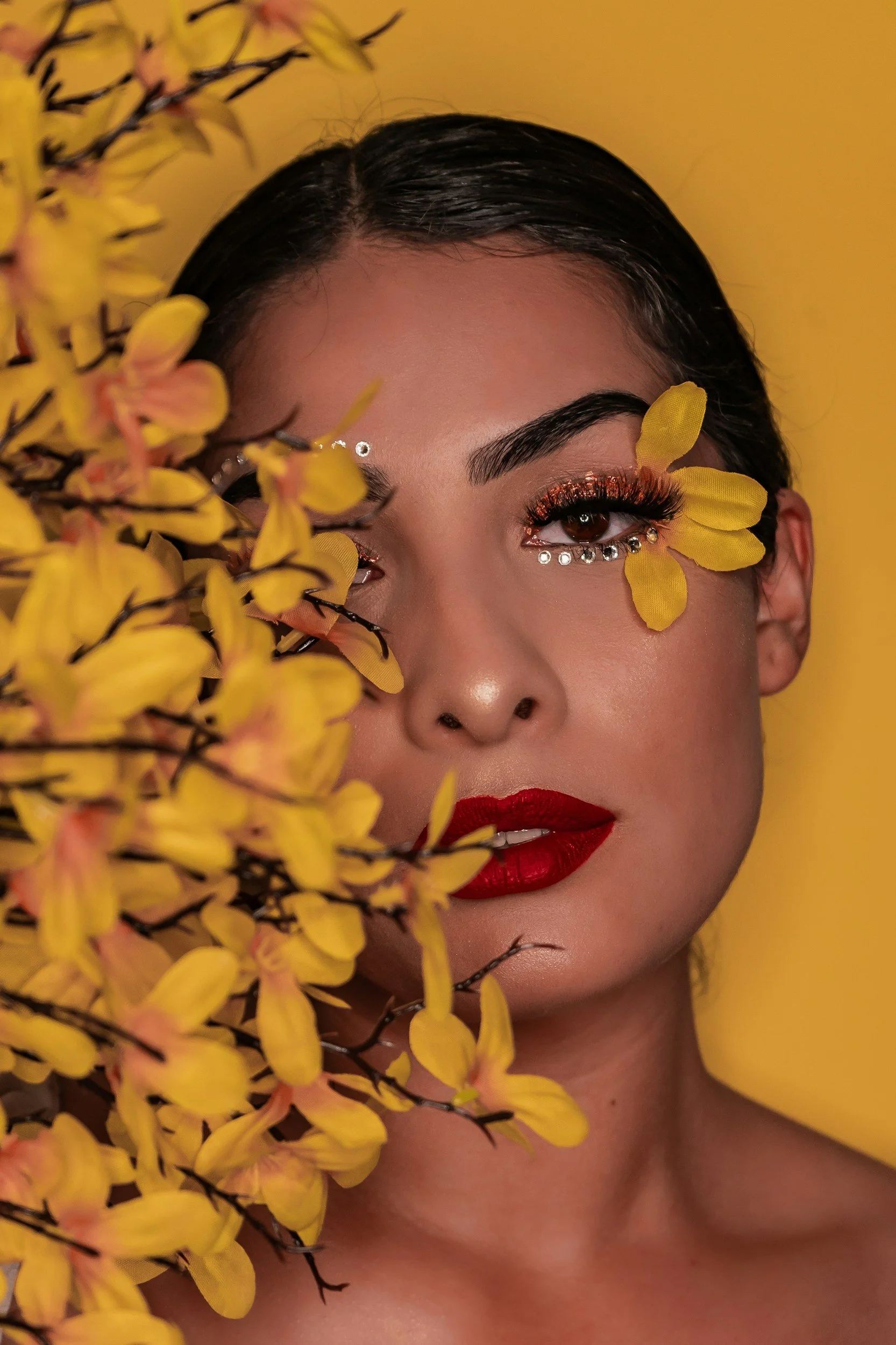 Close-up of a woman with dark hair, dramatic makeup with glittery orange eyeshadow, rhinestones under her eye, and bold red lipstick. She is partially obscured by yellow flower petals in the foreground, with a yellow background.