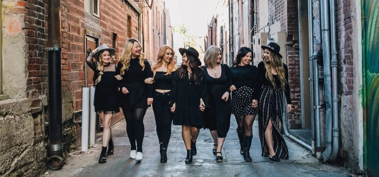 Group of eight women walking together down a narrow alley, smiling and holding hands, wearing stylish black outfits and hats.