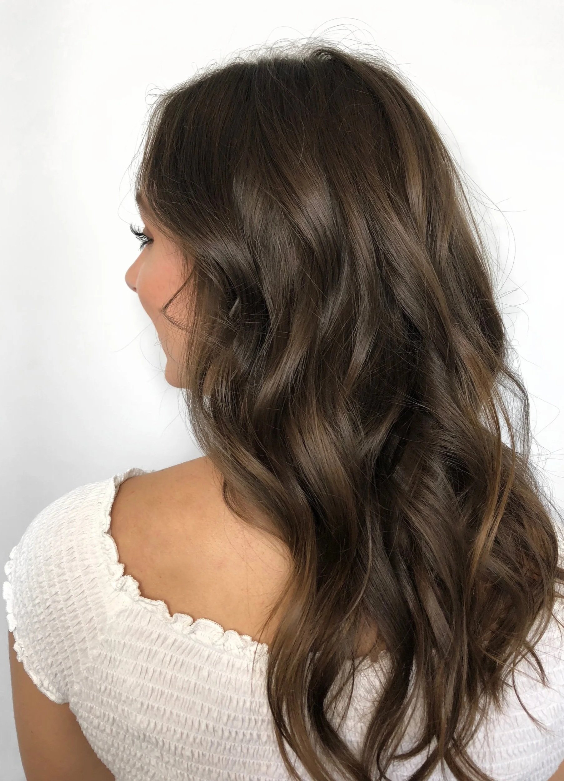 A woman with long, wavy brown hair seen from the back and side, wearing a white, textured short-sleeve top.
