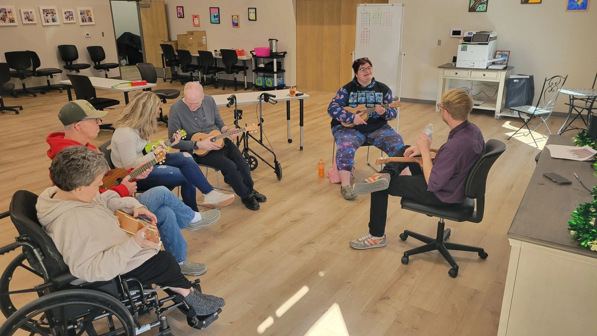 People playing ukuleles in a room, including a woman in colorful pajamas singing and playing ukulele, and several others seated with ukuleles. The room has a wooden floor, chairs, and artwork on the walls.