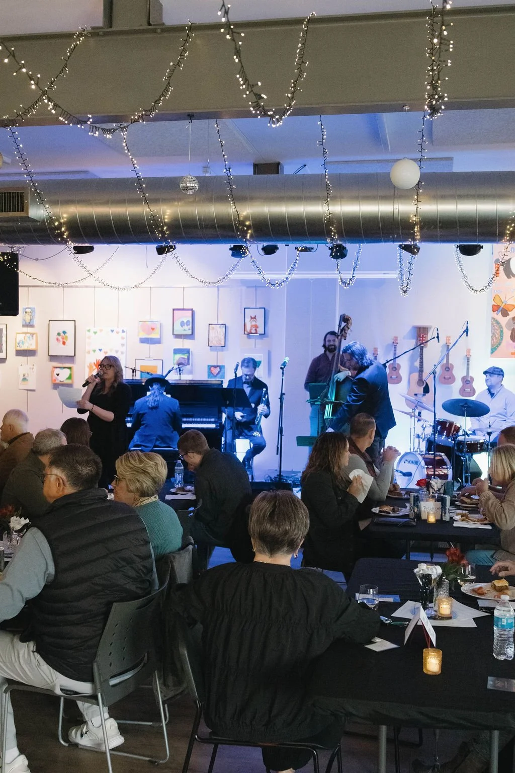 Live musical performance at an indoor event with decorated ceiling lights, audience seated at tables, and artwork on the wall.