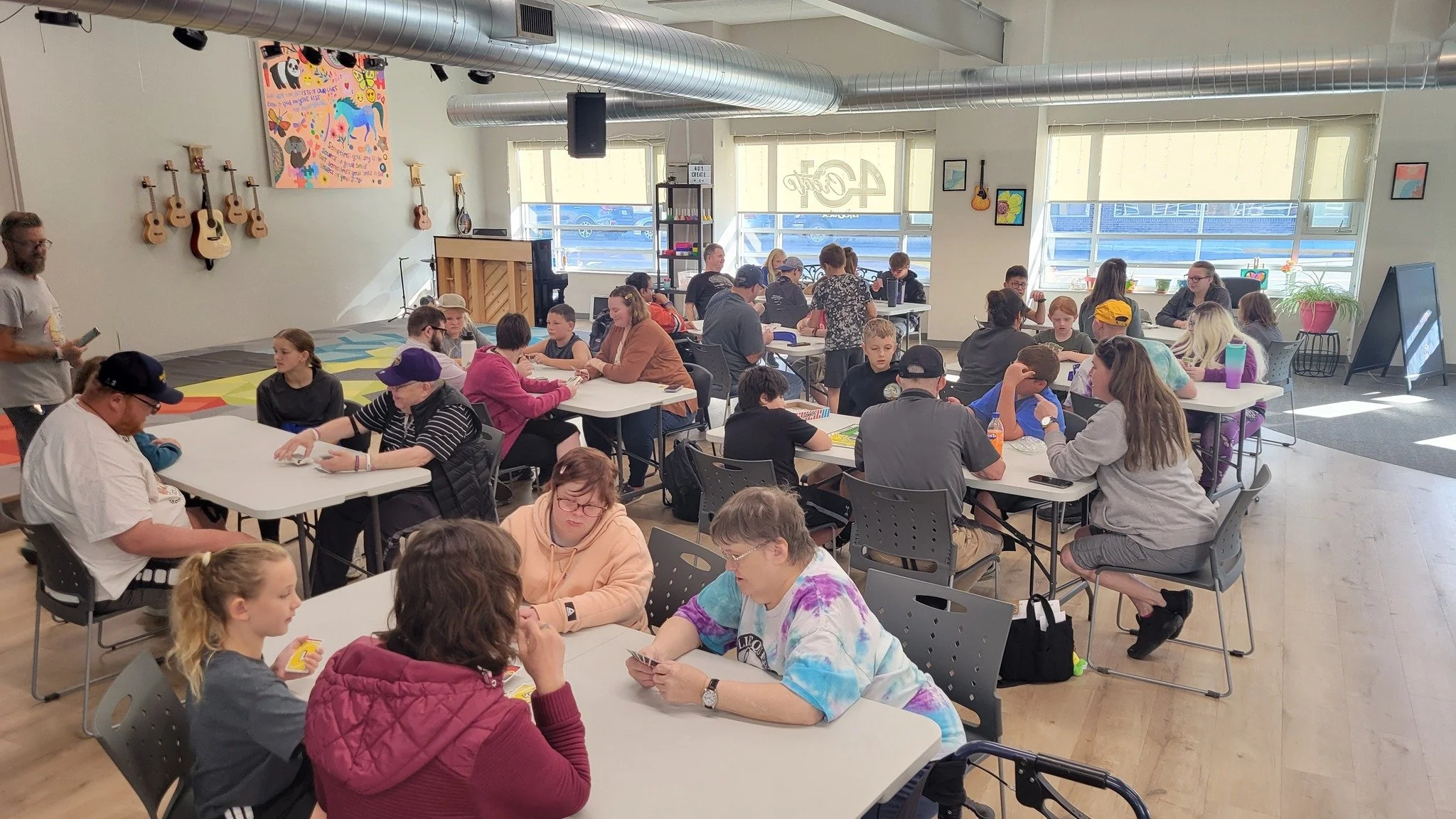 A large indoor gathering of people, mostly children and some adults, seated at tables engaged in various activities, with guitars hanging on the wall and colorful artwork on the walls.
