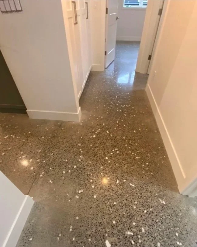 A hallway with white walls and baseboards, leading to a room with a window. The floor is polished concrete with white paint splatter.