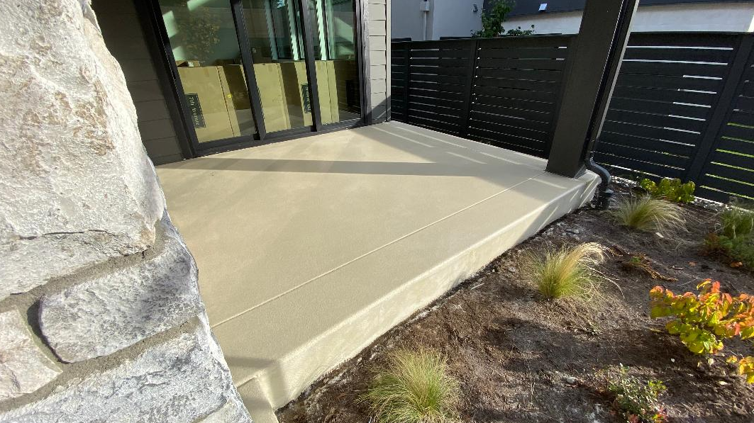 Modern outdoor patio with a concrete slab, black wooden fence, and surrounding plantings.